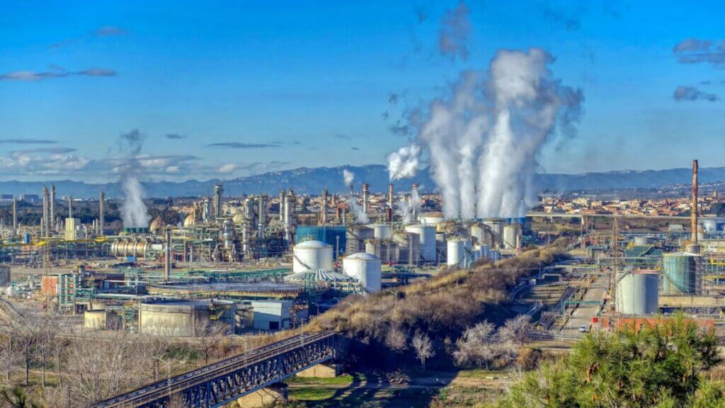 A large oil refinery complex with steam rising from multiple stacks under a clear blue sky.