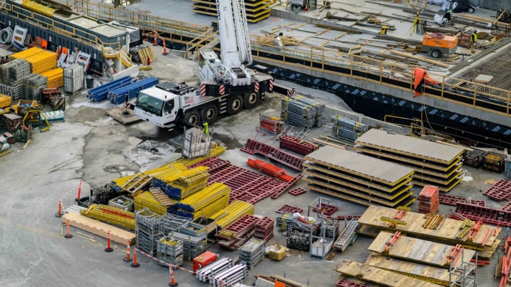 Aerial view of a construction site featuring stacked materials, orange cones, scaffolding, and a large crane.