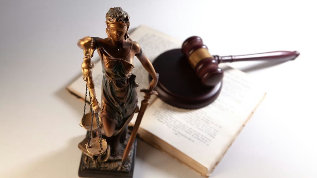 A bronze statue of Lady Justice holding scales and a sword, resting on an open book next to a dark wooden gavel.