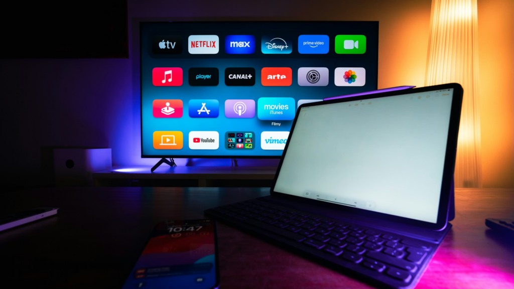 A dark room showing an Apple TV interface on a large screen with various streaming apps, with an iPad and smartphone in the foreground.