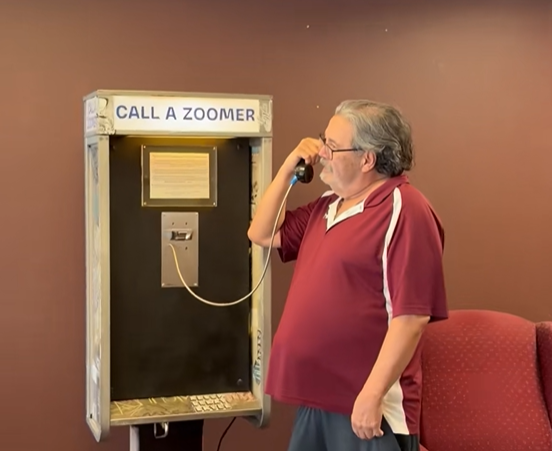 Older man using a “Call a Zoomer” payphone, holding receiver and speaking into handset indoors.