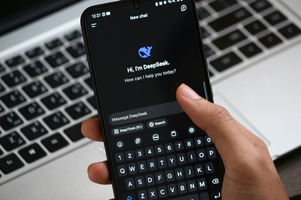Person holding smartphone showing DeepSeek chatbot interface with text input over a laptop keyboard.