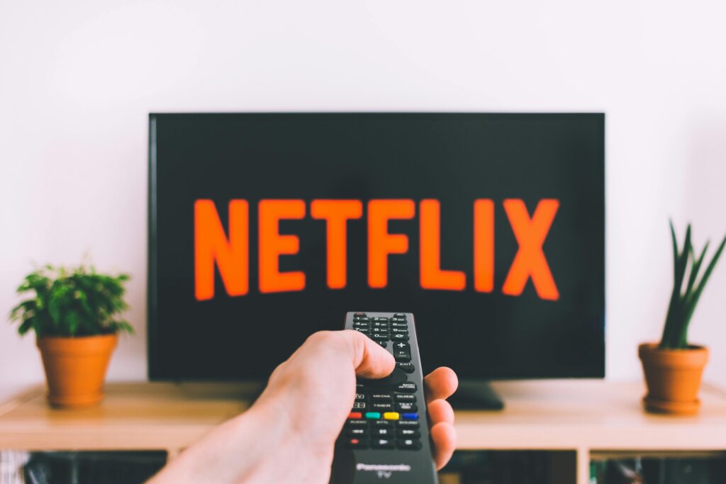 Hand holding a remote control pointing at a TV screen displaying the Netflix logo.