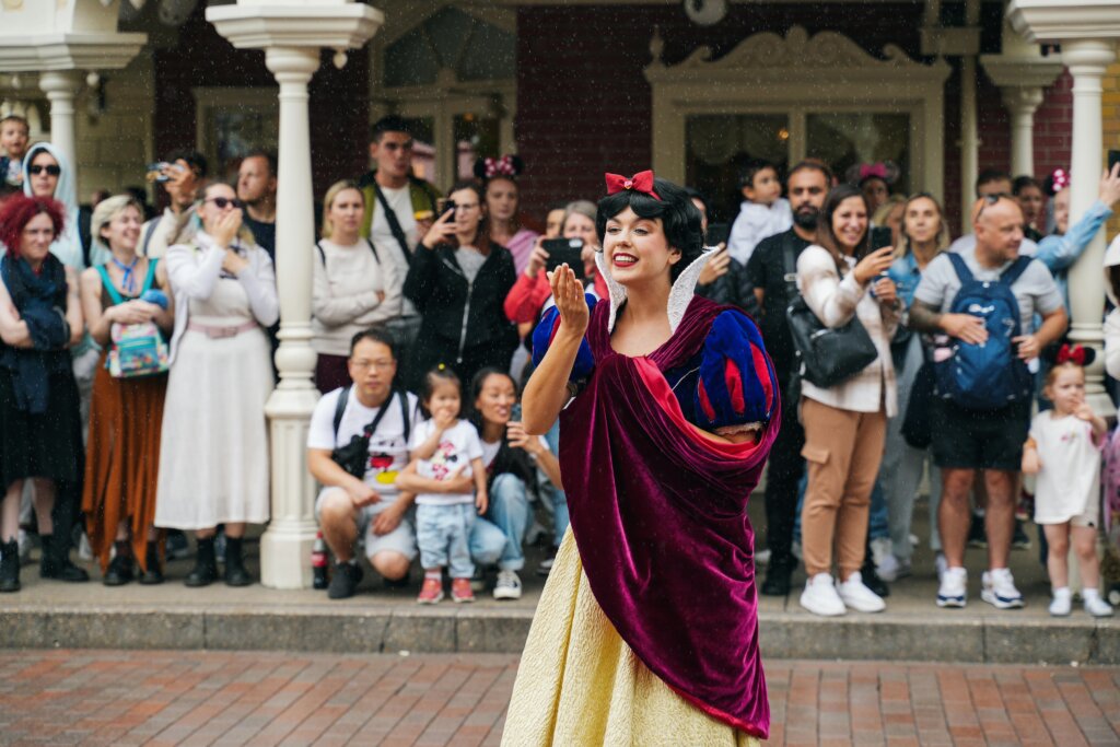 Snow White performer blowing a kiss to crowd during Disney park parade as visitors watch and take photos.