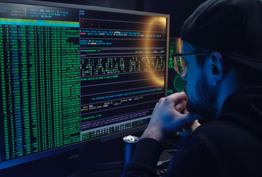 Man wearing glasses analyzing code on multiple monitors in a dimly lit workspace.