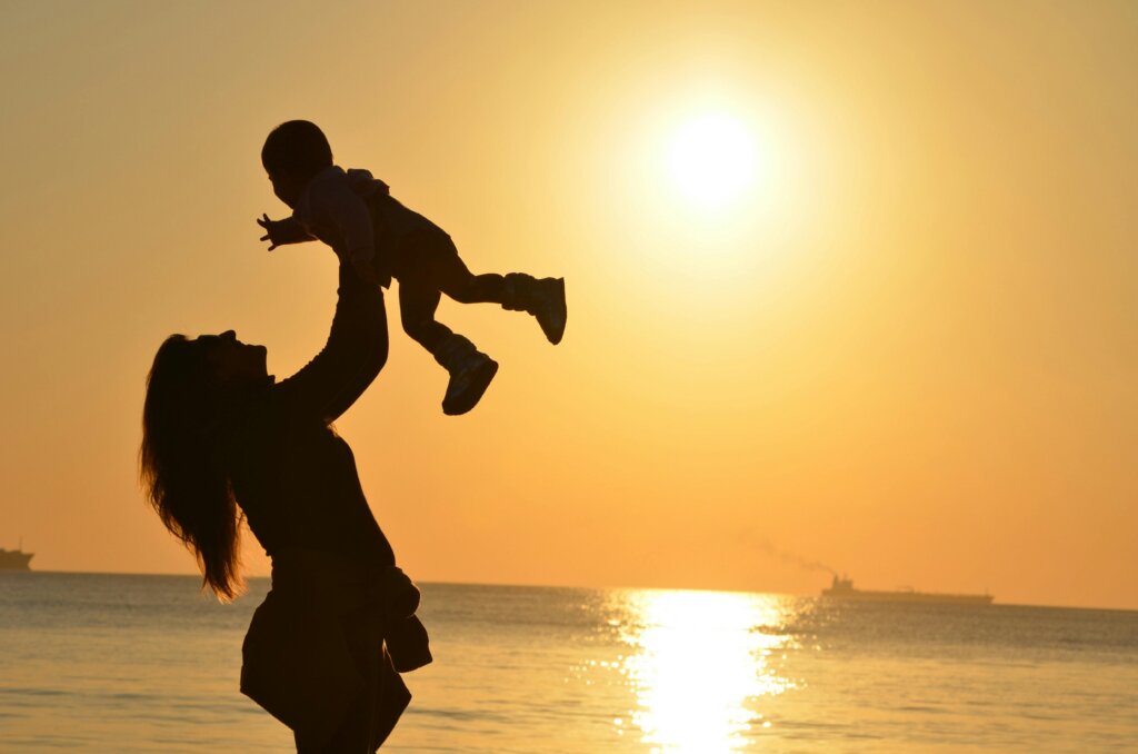 Silhouette of a woman lifting a baby in the air at sunset by the ocean.