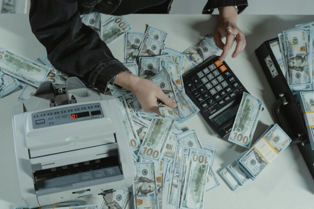 Person counting stacks of US dollar bills beside a calculator and cash counting machine on a desk.