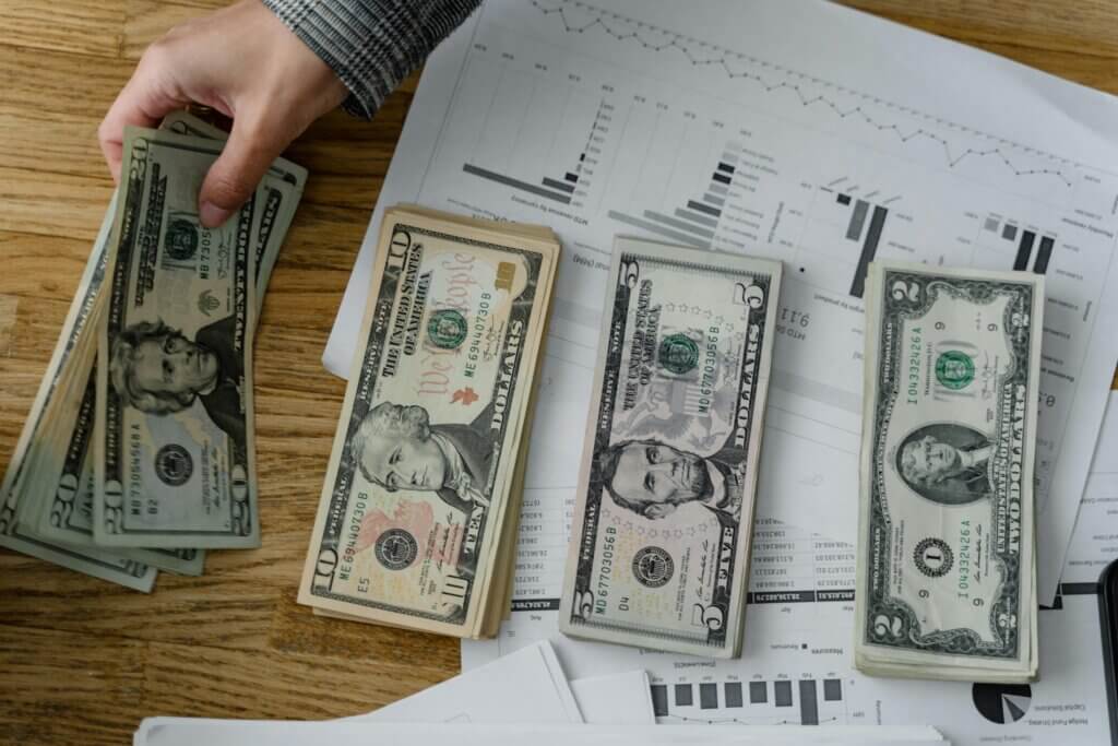 Stacks of US currency arranged on a table with financial charts and a hand placing additional bills.