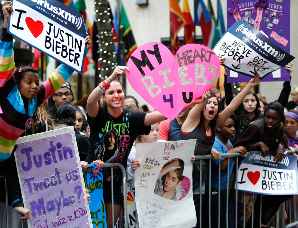 Justin Bieber fans holding signs and heart-shaped posters while gathered behind barricades