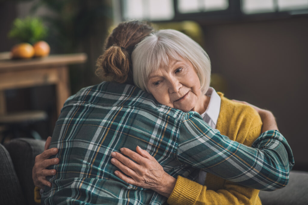 Older woman hugging a younger person in a living room, resting her head on their shoulder.