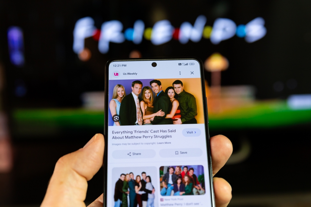 Smartphone displaying a Friends cast photo on screen with a blurred Friends sign in the background.