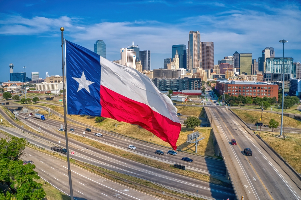 Texas state flag waving above a highway with the Dallas skyline in the background.