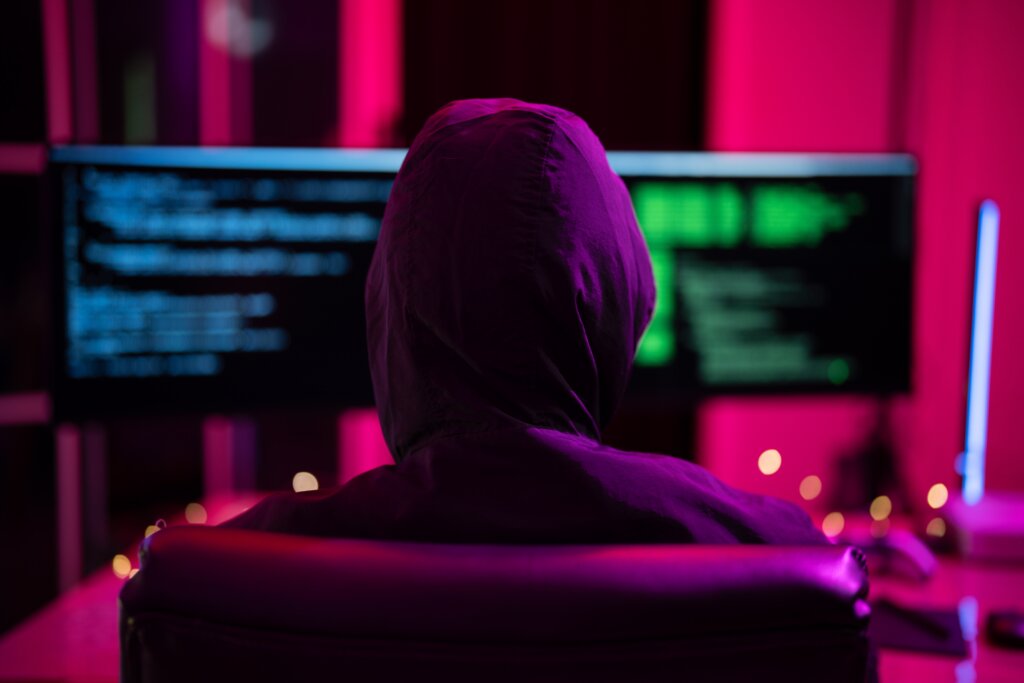 Person in a hoodie facing computer screens displaying code in a dark, neon-lit room.