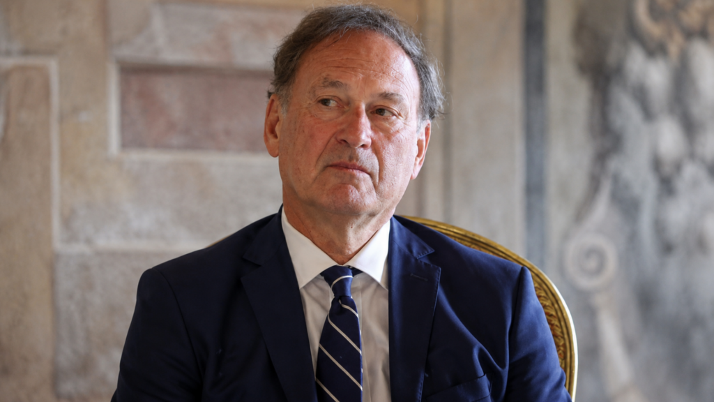 A close-up, slightly low-angle portrait of Justice Samuel Alito looking thoughtfully toward the side; he is wearing a navy blue suit and a diagonally striped tie, set against an ornate, blurred interior background.