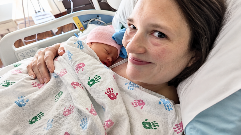 A close-up, heartwarming shot of a smiling woman lying in a hospital bed, cradling her newborn baby; the mother is wearing a white hospital gown decorated with colorful hand-prints, while the sleeping infant wears a small pink cap.