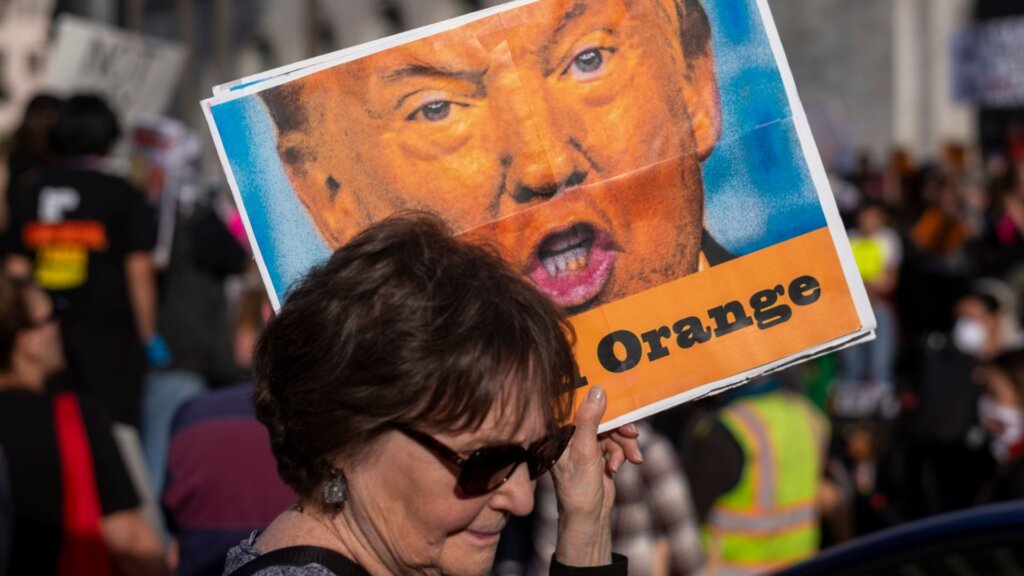 Woman wearing sunglasses at a crowded protest holds a large poster showing a close up caricature of Donald Trump with the word “Orange” printed in bold black letters across the bottom.