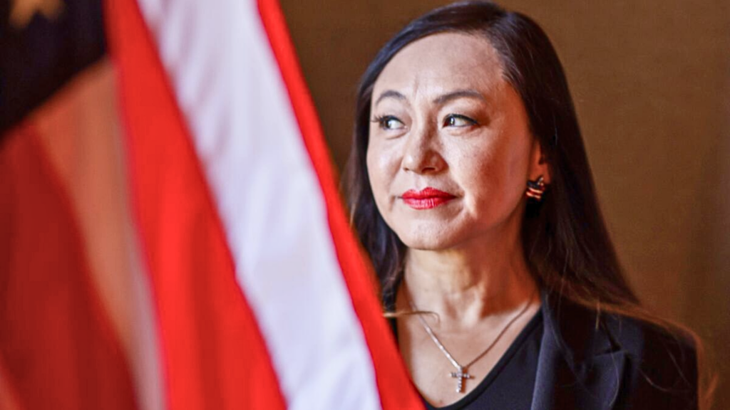A close-up portrait of former Mayor of Arcadia Eileen Wang, she is looking slightly off-camera with a gentle smile, wearing a dark top, a cross necklace, and American flag earrings, with part of a U.S. flag visible in the foreground.