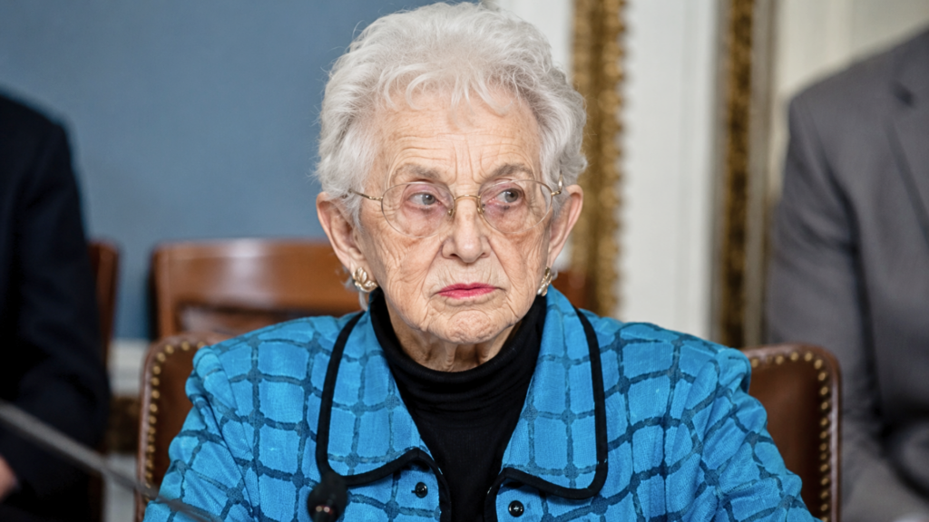 A close-up portrait of Representative Virginia Foxx looking thoughtfully to the side while resting her chin on her hand; she is wearing a vibrant blue-and-black patterned jacket, a black turtleneck, and gold-rimmed glasses, with a blurred American flag in the background.