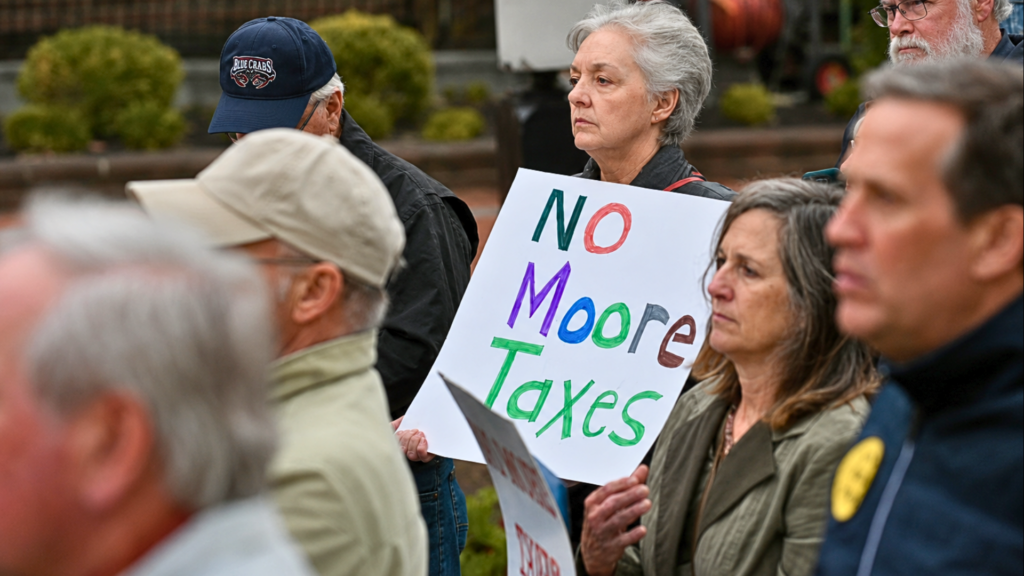 A close-up shot focused on a group of local citizens standing outdoors; an older woman with short grey hair and glasses looks forward intently, holding a handwritten white cardboard sign that reads "NO Moore Taxes" in multi-colored block lettering, capturing an active tax protest.