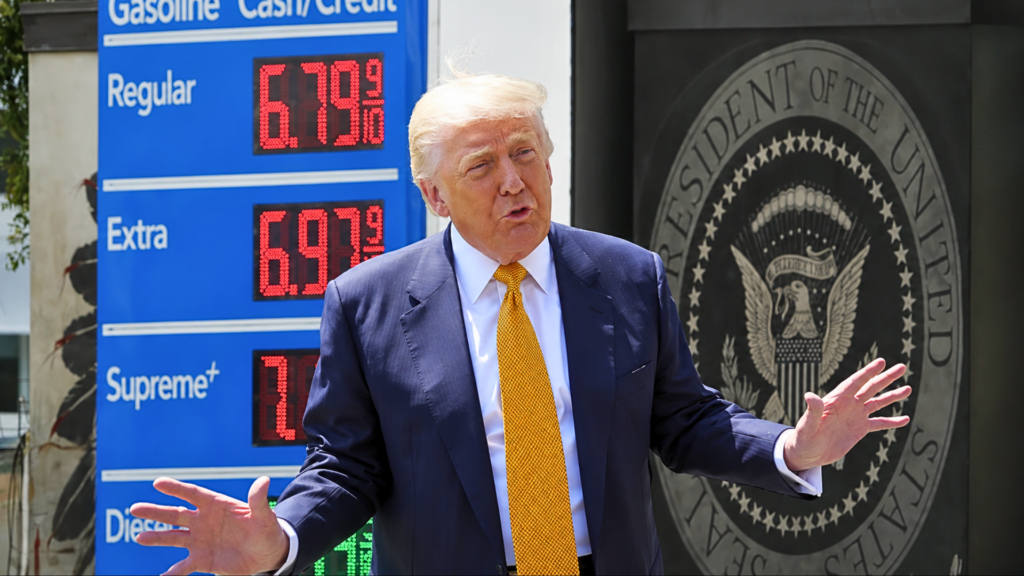 A close-up shot of Donald Trump speaking expressively with both hands raised; he is wearing a dark blue suit with a bright yellow tie, positioned between a blue gas station price sign reading 6.79 9/10 and a black backdrop featuring the seal of the President of the United States.
