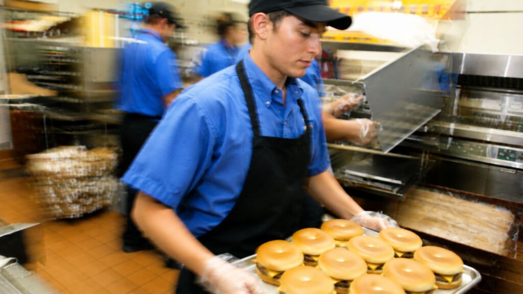 Fast food worker in a blue uniform carries a tray of cheeseburgers through a busy restaurant kitchen while coworkers prepare food in the background.