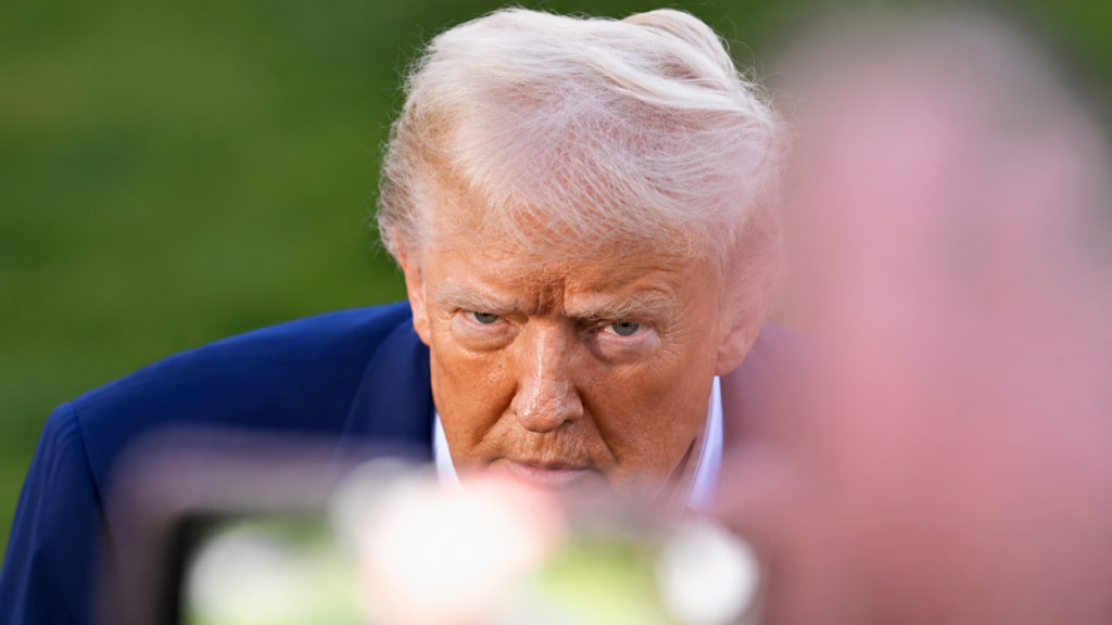 A dramatic, close-up portrait of President Donald Trump looking sharply forward with a stern, intense glare; his face is tightly framed between a blurry cell phone camera in the foreground and a rich green grass background.