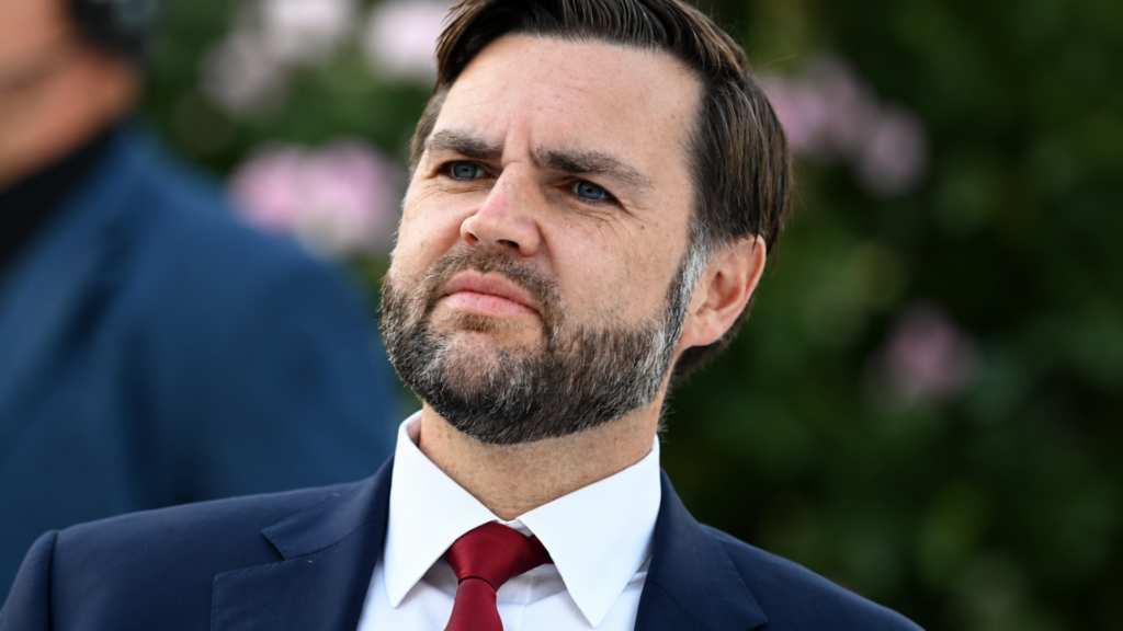 A close-up, slightly low-angle portrait of Vice President JD Vance looking off-camera with a stern, focused expression; he is wearing a dark blue suit and a solid crimson tie, captured against a soft, out-of-focus background of green foliage.