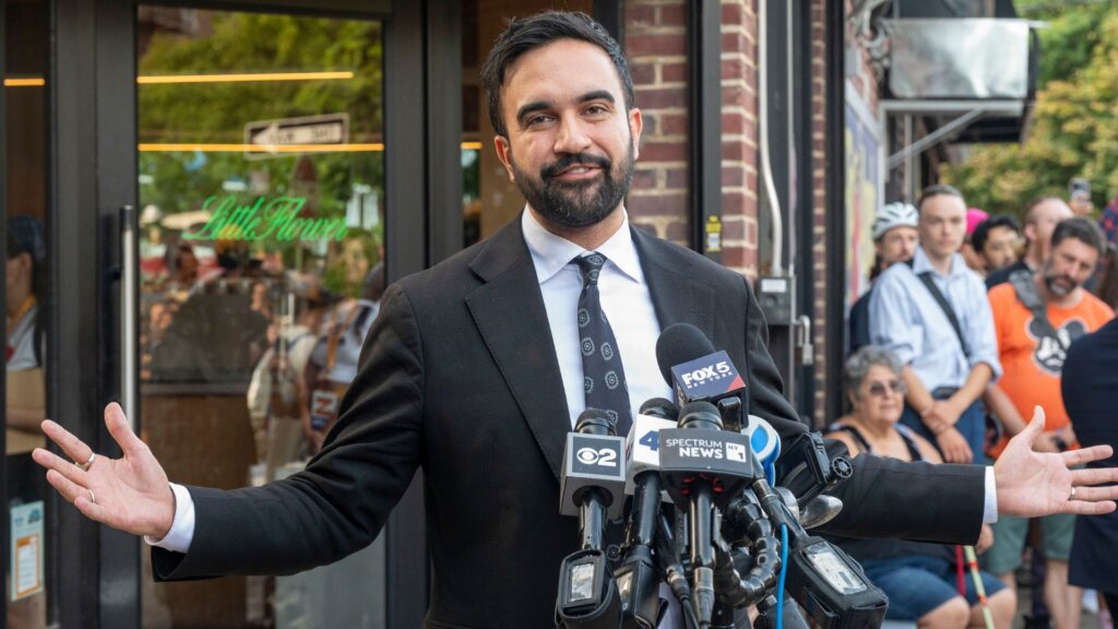 New York City Mayor Zohran Mamdani, in a dark suit speaking to reporters outside a storefront while standing in front of several news microphones.