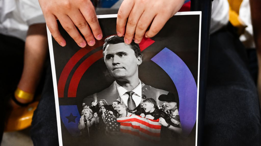 A close-up shot of hands holding a political flyer featuring a portrait of conservative activist Charlie Kirk surrounded by supporters and an American flag.