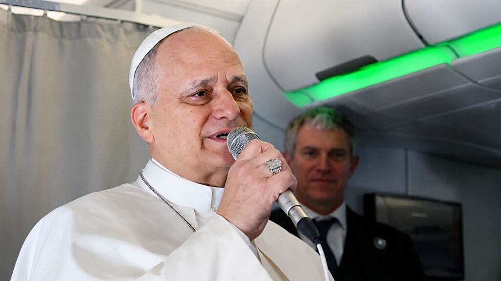 Pope Leo speaks into a microphone while standing inside an airplane cabin, wearing white papal robes and a skullcap.