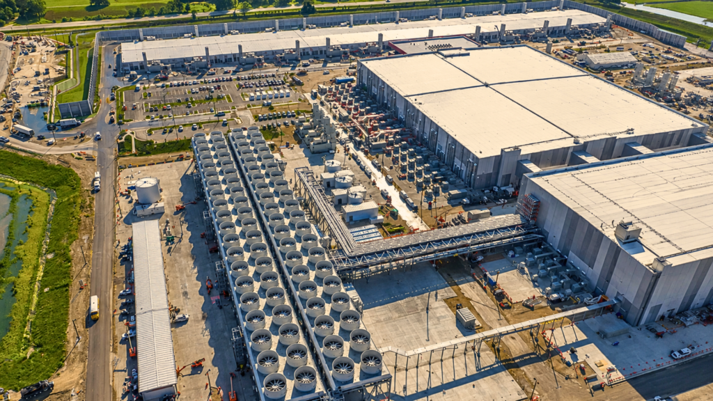 An aerial view of a large data center complex featuring an expansive bank of white cooling fans, electrical substations, and multi-story server buildings.