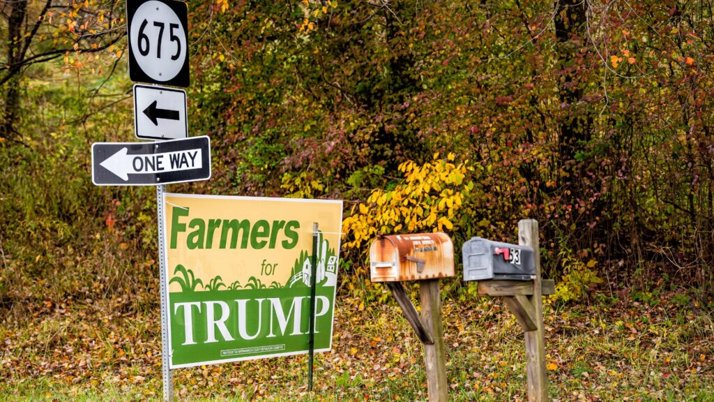 Rural roadside scene in autumn with a campaign sign reading “Farmers for TRUMP” posted beneath highway marker “675” and a “ONE WAY” sign. Mailboxes stand beside the sign against a backdrop of colorful fall foliage.