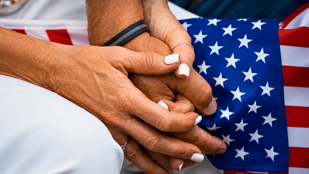 A close-up, high-angle shot of two pairs of hands clasping together over an American flag; the skin is weathered, representing an older generation, and one person wears a thin silver ring while the other wears a black-and-grey silicone band.