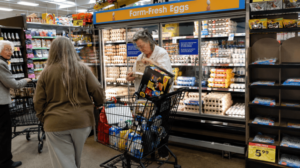 An older woman selects a carton of eggs from a fully stocked refrigerated display case under a "Farm-Fresh Eggs" sign in a grocery store aisle.