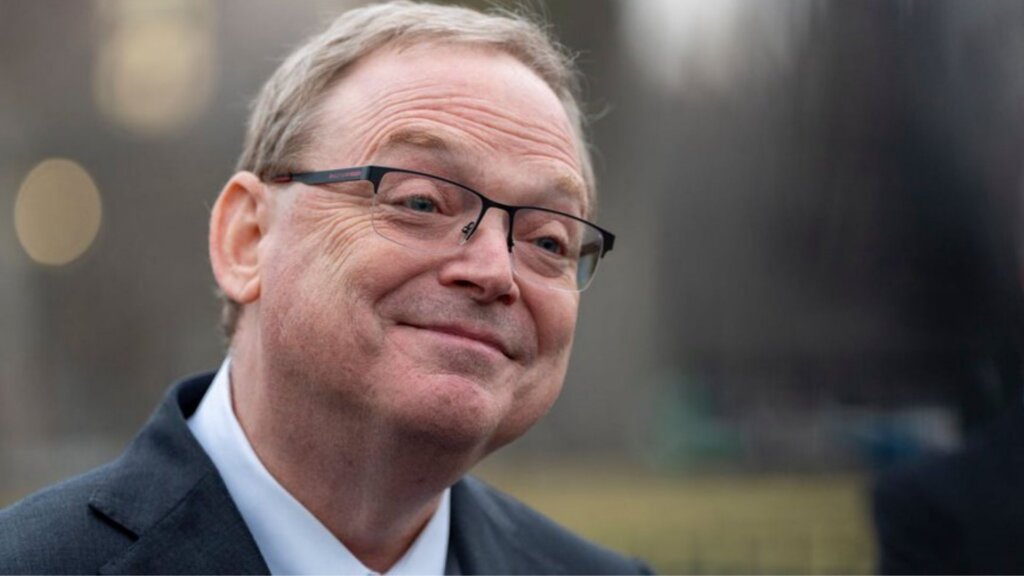 Close-up portrait of Kevin Hassett wearing glasses and a dark suit jacket, smiling slightly while looking off to the side outdoors with a blurred city background behind him.