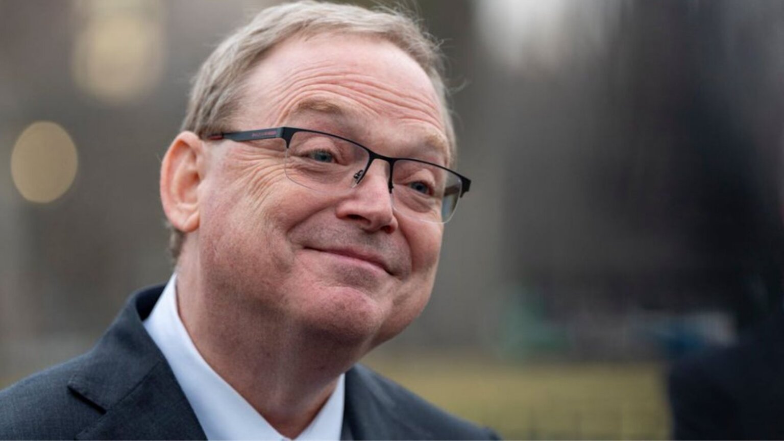 Close-up portrait of Kevin Hassett wearing glasses and a dark suit jacket, smiling slightly while looking off to the side outdoors with a blurred city background behind him.