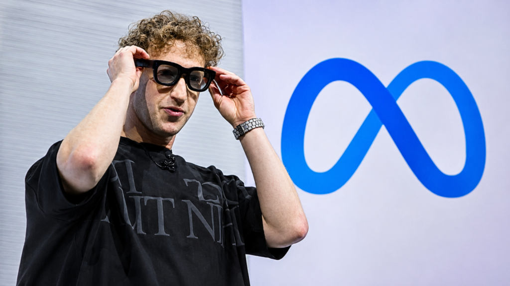 A medium shot of Meta CEO Mark Zuckerberg looking thoughtfully to the side while adjusting a pair of chunky, black-framed Orion AR glasses on his face; he is wearing an oversized black t-shirt, with the bright blue Meta logo visible in the background.