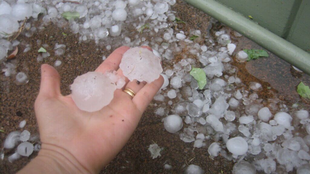 Hand holding large hailstones next to scattered ice on the ground after a storm, showing property damage often linked to insurance claims.