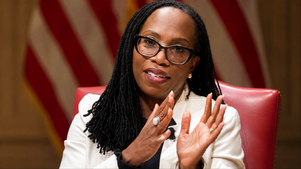 A medium shot of Justice Ketanji Brown Jackson speaking expressively with both hands raised; she is wearing a white blazer over a black top and her signature black-rimmed glasses, with a blurred American flag behind her.