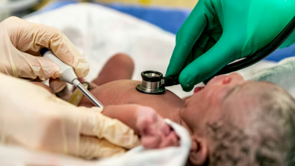 A close-up medical shot of a newborn's chest being examined; a healthcare worker in a green glove holds a stethoscope to the baby's heart, while another hand in a tan glove holds a medical probe, illustrating a neonatal cardiac evaluation.