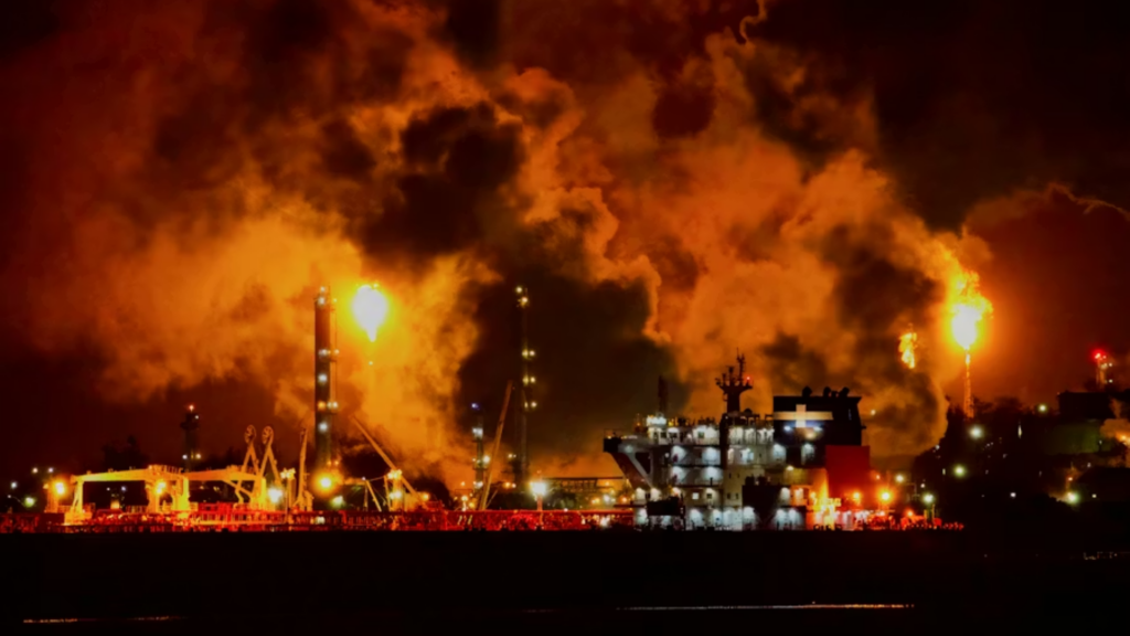 A dramatic nighttime shot of an industrial oil refinery complex ablaze with orange light; thick plumes of smoke rise into the dark sky as vertical gas flares emit massive balls of fire next to a production vessel docked along the water.