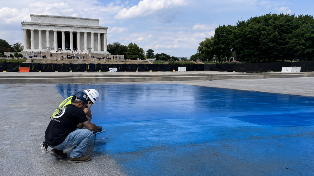 A wide exterior shot of the National Mall showing two construction workers in hard hats and safety vests kneeling on the stone basin of the Lincoln Memorial Reflecting Pool; they are applying a smooth, vibrant layer of bright blue coating to the concrete floor, with the grand marble columns of the Lincoln Memorial standing prominently in the background under a blue sky.