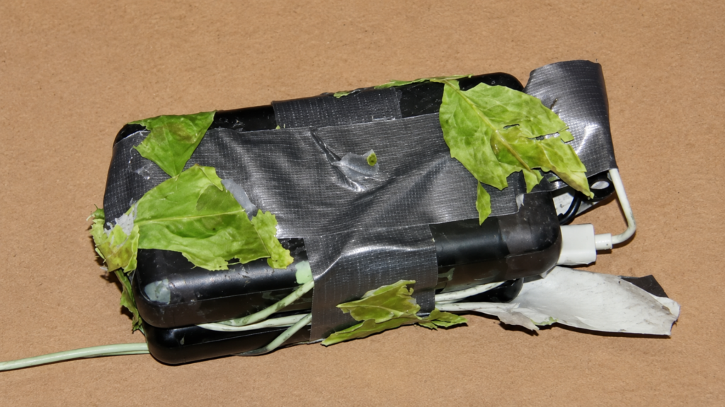 A close-up studio shot of a captured surveillance device; a black rectangular power bank and wiring are wrapped tightly in silver duct tape, with several green leaves pressed onto the exterior as crude camouflage over a white USB cable.