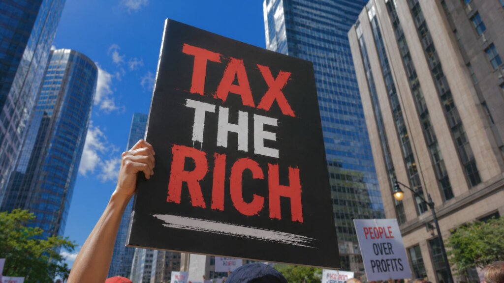 Protest sign reading “TAX THE RICH” held above a crowd during a city demonstration surrounded by tall downtown skyscrapers under a bright blue sky.