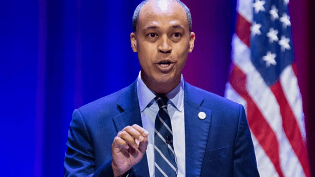 A medium shot of Virginia Attorney General Jay Jones speaking at a podium; he is wearing a sharp navy blue suit and a diagonally striped tie, gesturing emphatically against a vibrant blue-and-red stage backdrop with a blurred American flag.