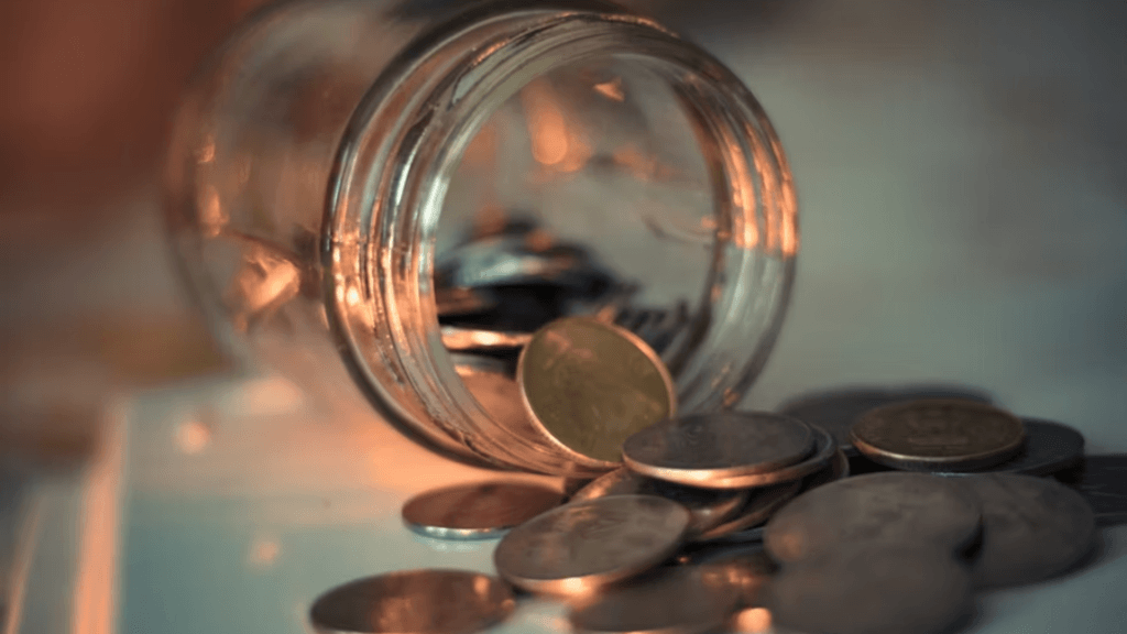 A close-up shot of an overturned glass jar with various metallic coins spilling out onto a light-colored surface.