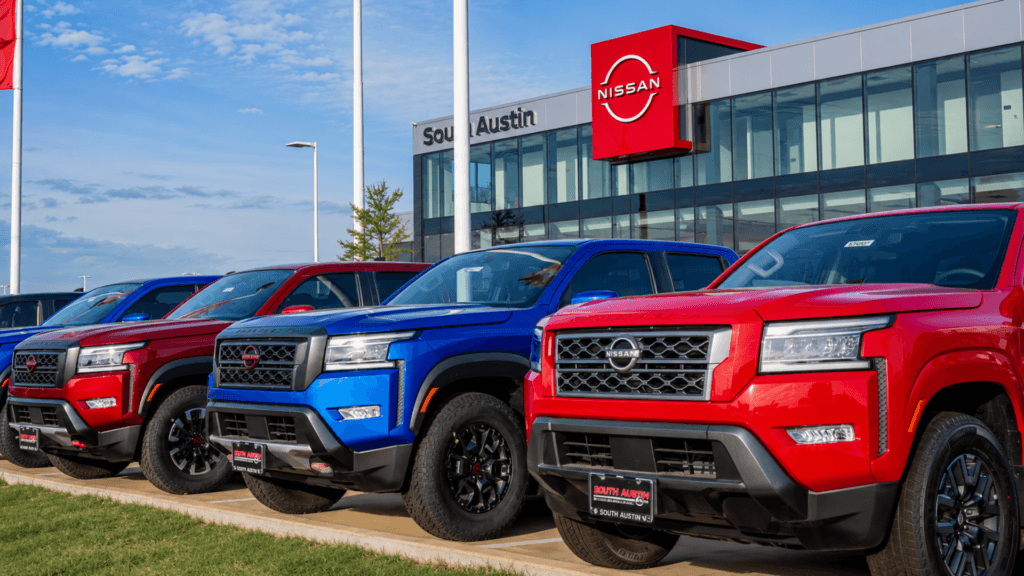 A wide daytime shot of several new Nissan Frontier pickup trucks in red and blue parked in a row at the "South Austin" Nissan dealership; the modern glass dealership building with a large red Nissan logo is visible under a clear blue sky.