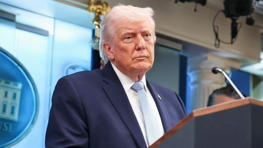 A medium shot of Donald Trump standing behind a dark wooden podium at the White House Press Briefing Room; he wears a dark blue blazer and a light blue textured tie, looking out with a serious, focused expression.