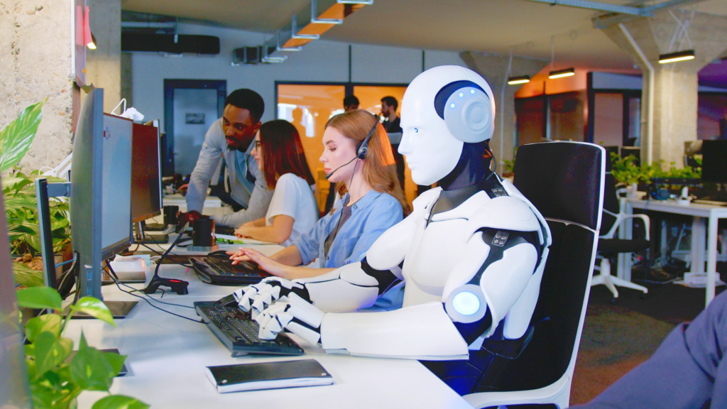 A humanoid robot sitting at a desk and typing on a keyboard in a busy, modern open-plan office alongside human workers.