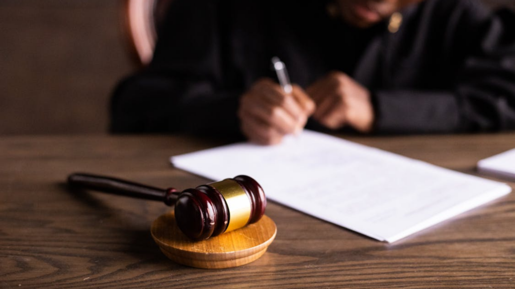 A wooden judge's gavel rests on a desk while a person in a black robe writes on a document in the blurred background.